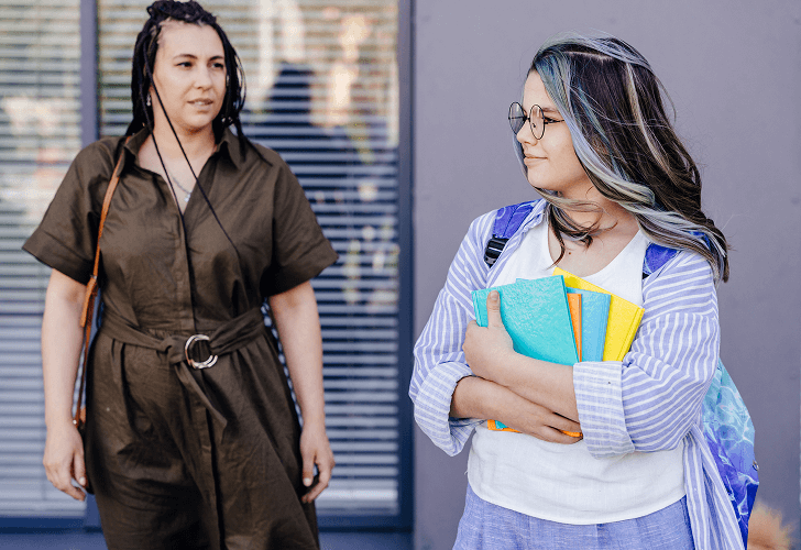 A student holding books, smiling slightly, while an adult stands nearby, observing and offering support.