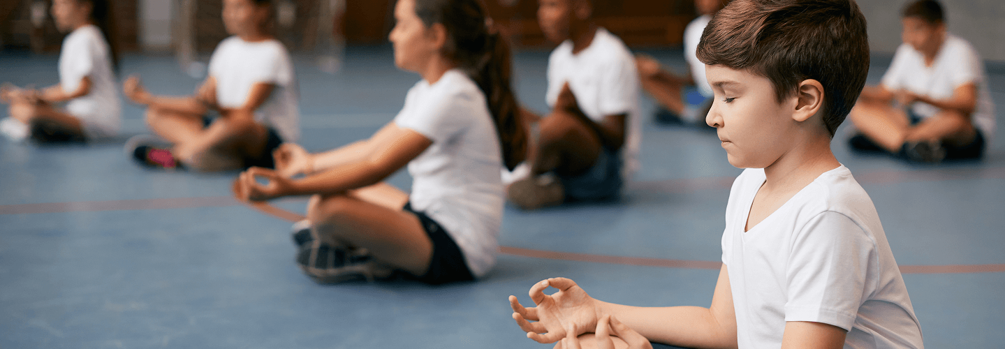 Group of children sitting cross-legged on the floor practicing mindfulness or meditation in a gymnasium.