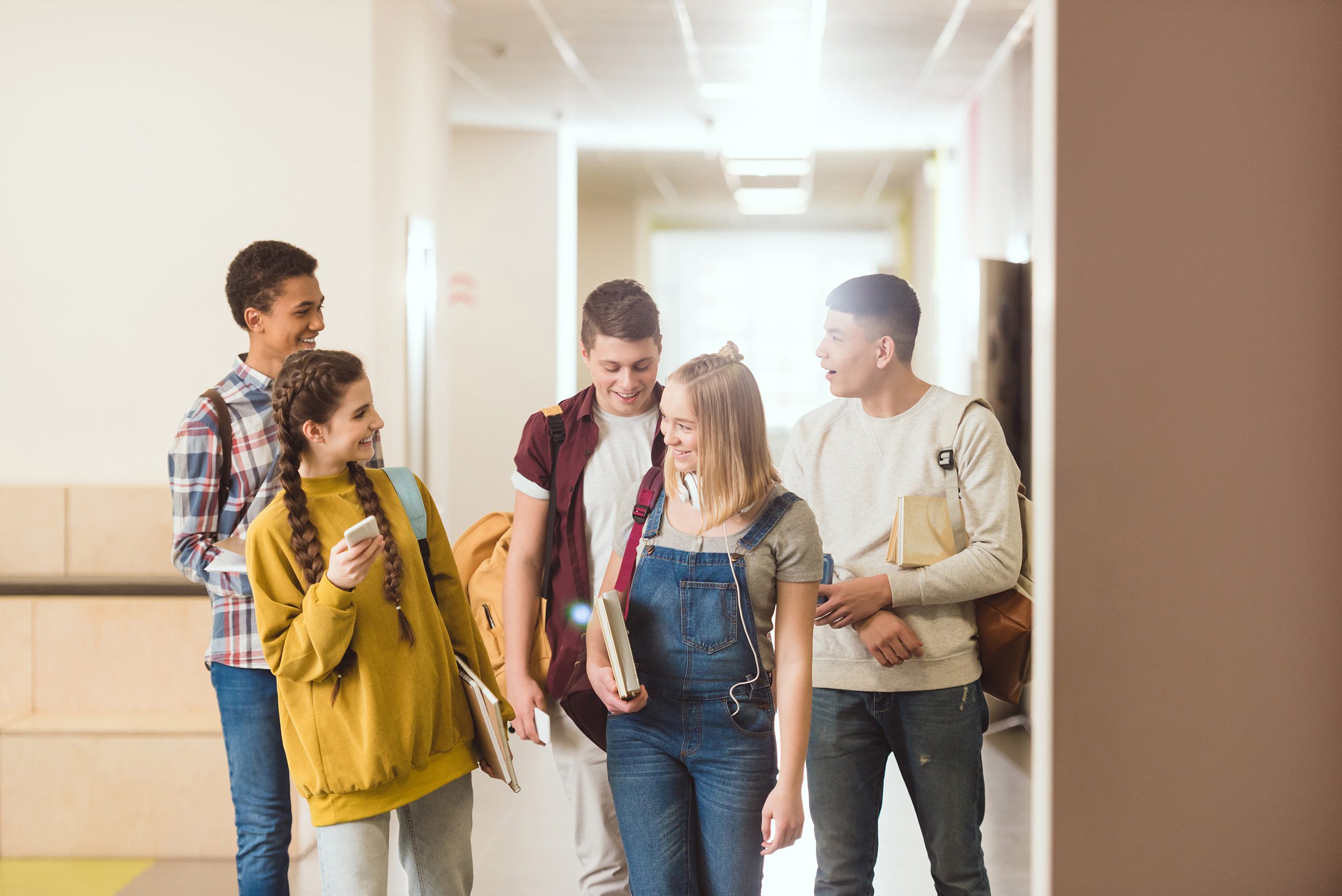 Group of five diverse high school students walking and talking together in a school hallway, smiling and carrying books and backpacks.